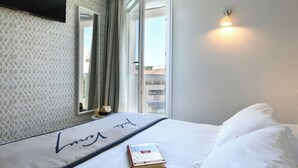 A hotel room with a bed, a book, a flat-screen TV, a mirror, and a window with curtains. - Hôtel Jules Verne Biarritz (Biarritz)