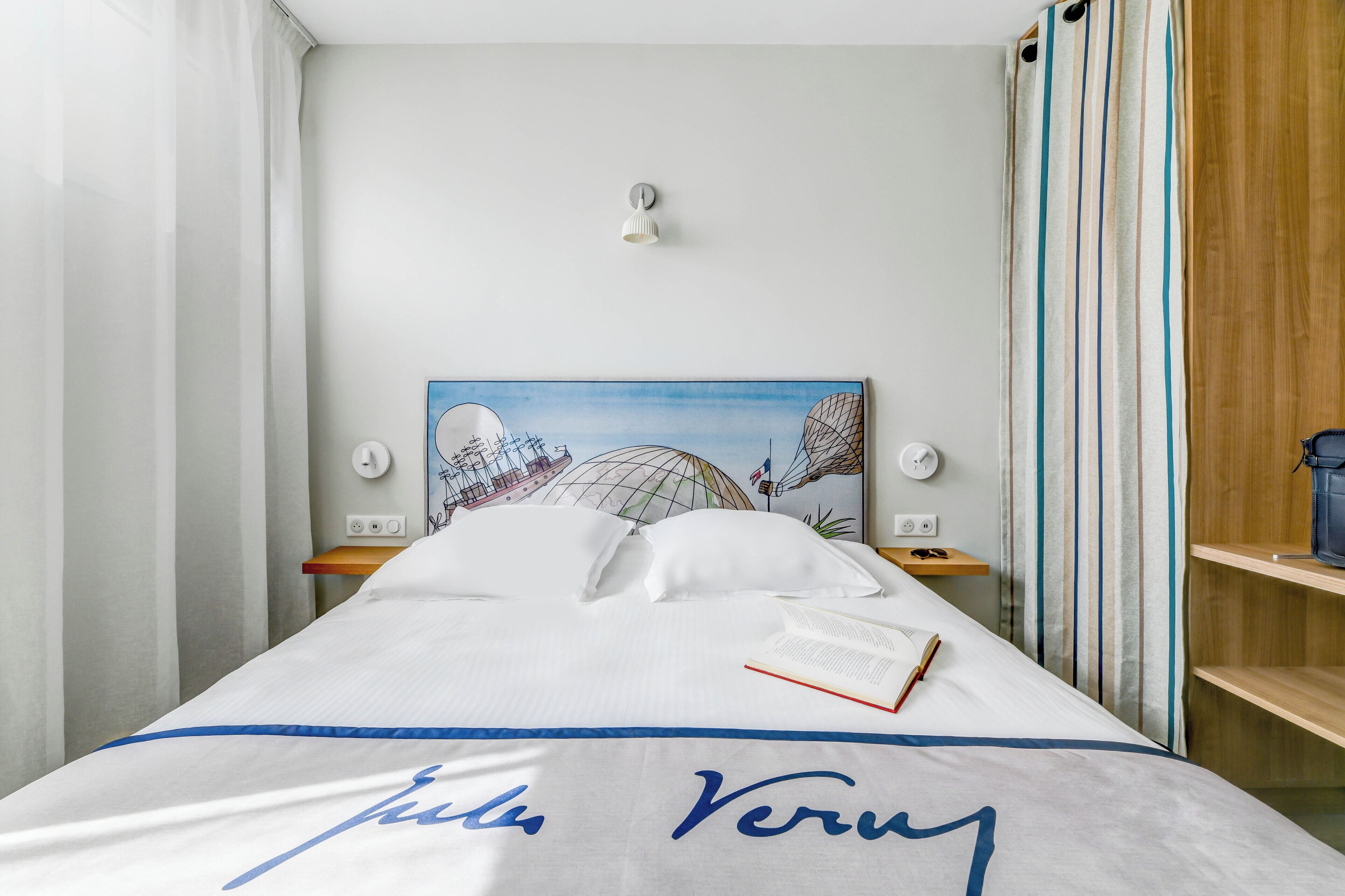 A neatly made bed with a white and blue bedspread, a book, and a lamp on a bedside table.