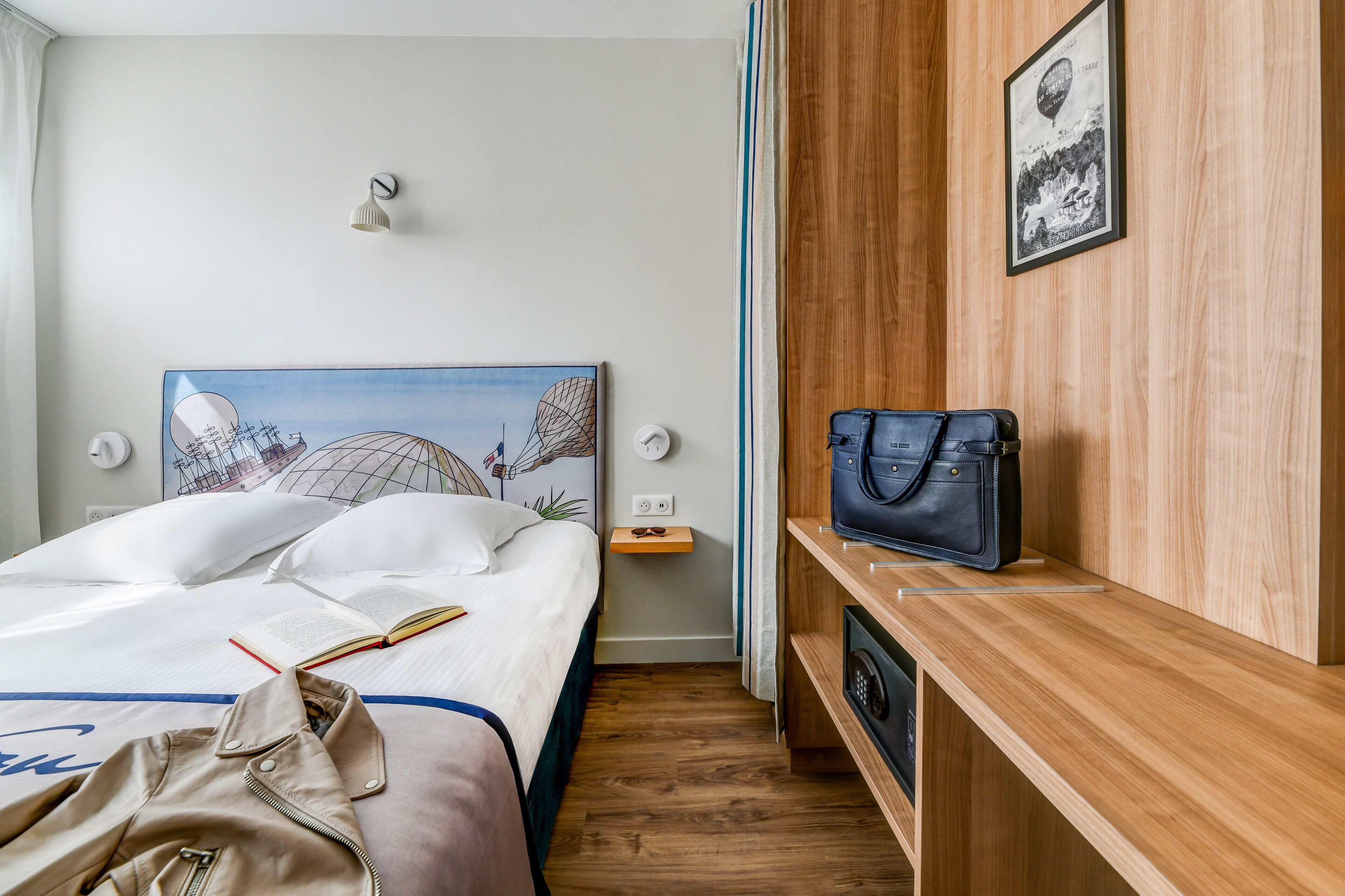 Un lit bien fait, avec une tête de lit représentant un paysage urbain, une table de chevet avec une lampe et une étagère en bois sur laquelle repose un sac à main.