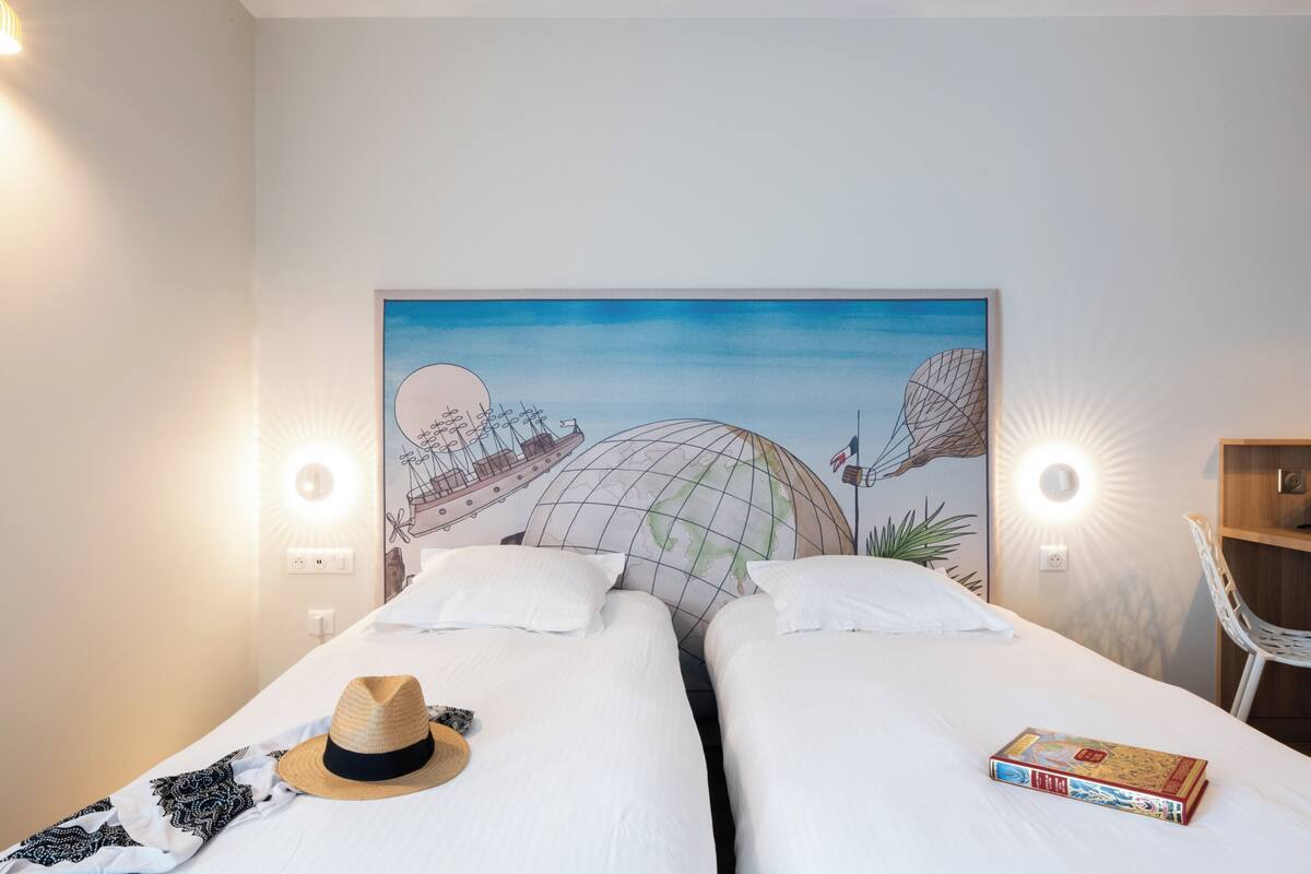 A hotel room with two beds, a globe artwork on the wall, a hat, and a book on one bed.