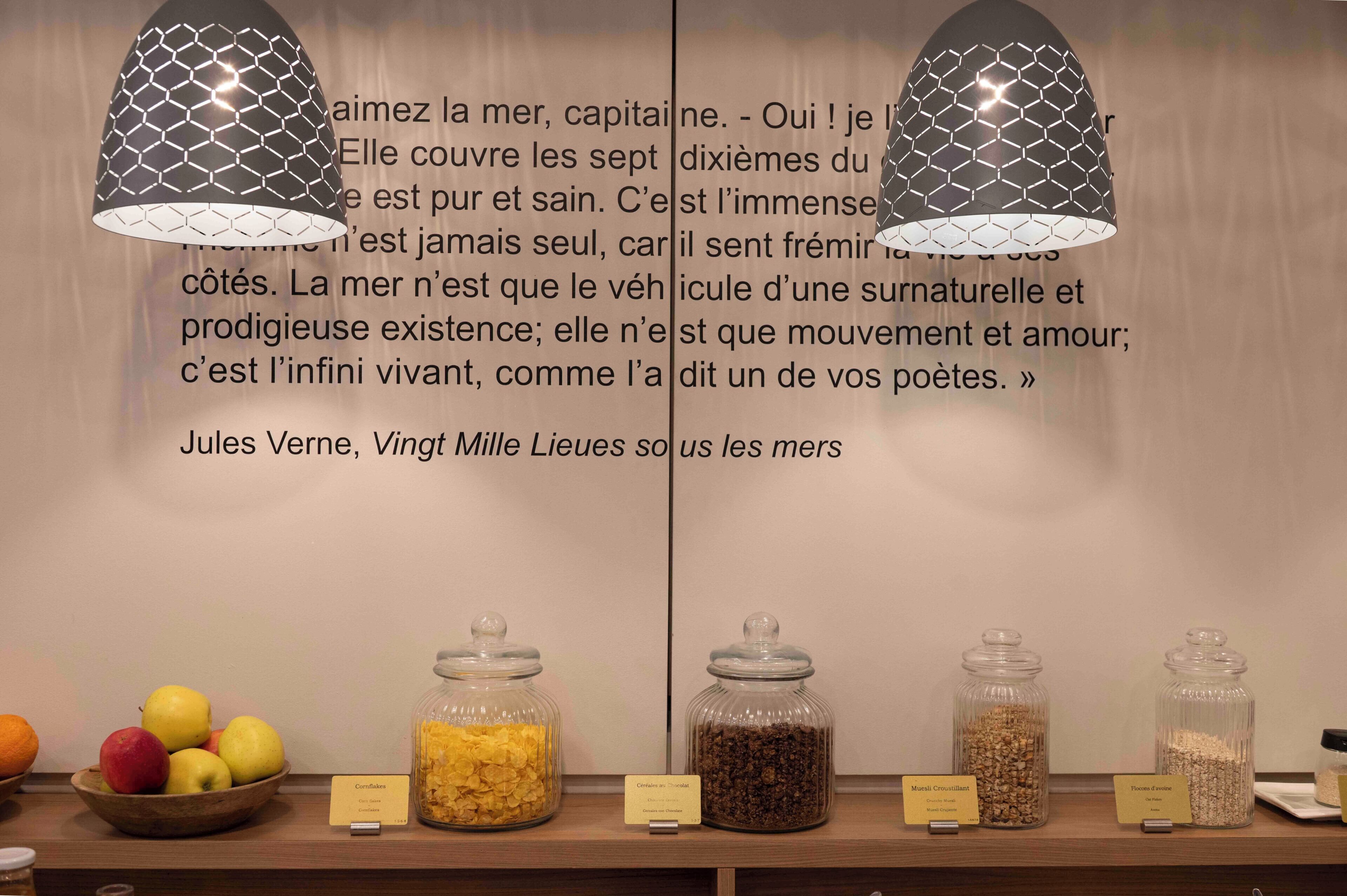 Una habitación con una pared que presenta una cita en francés, una mesa de madera con fruta y frascos de comida etiquetados, y dos lámparas colgantes con un diseño geométrico.