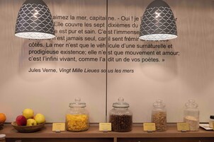 Una habitación con una pared que presenta una cita en francés, una mesa de madera con fruta y frascos de comida etiquetados, y dos lámparas colgantes con un diseño geométrico.