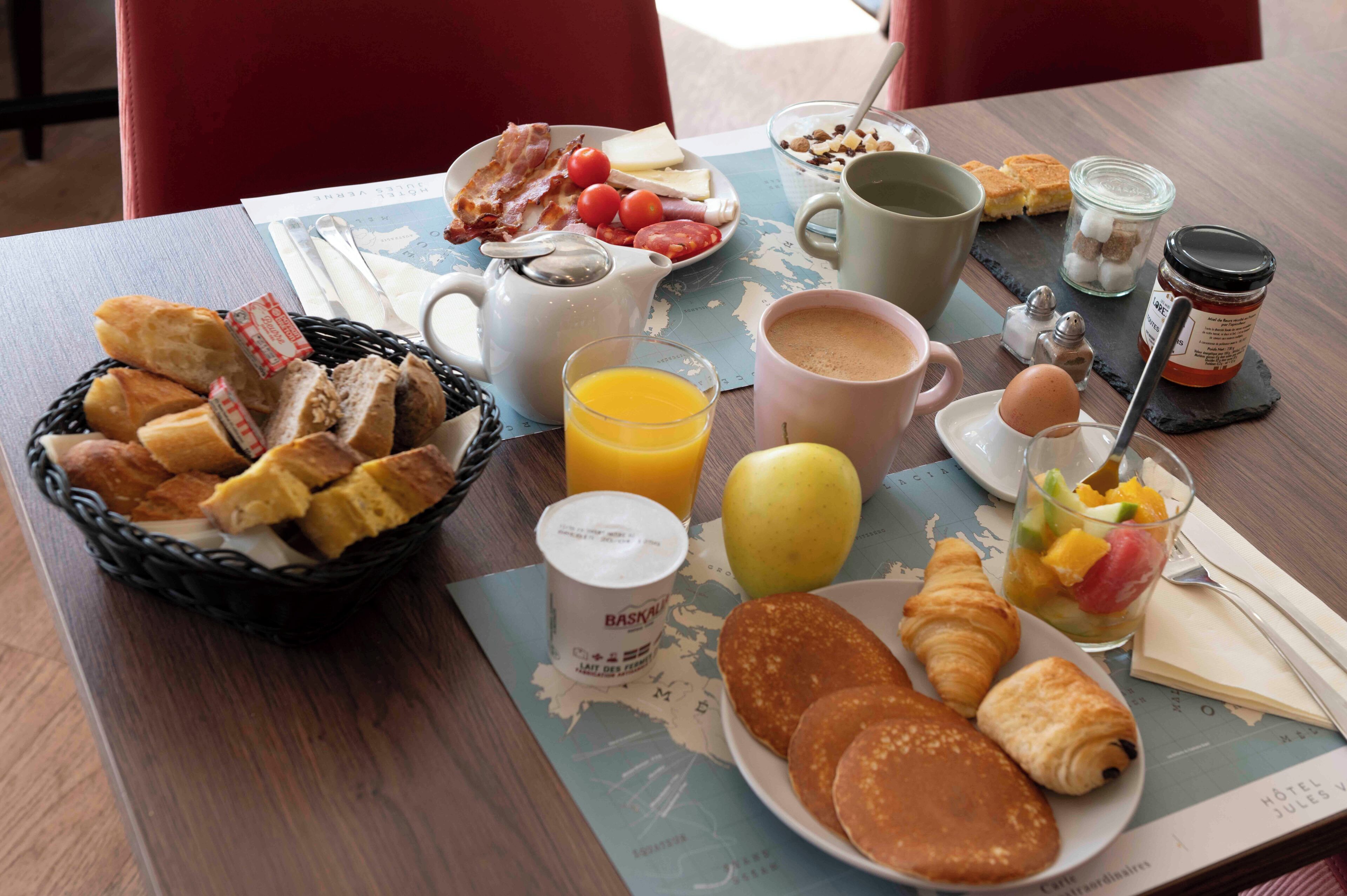 Una mesa de desayuno con croissants, panqueques, huevos, un frutero, una taza de café, un vaso de jugo y una cesta de pan.
