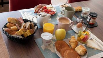 Una mesa de desayuno con croissants, panqueques, huevos, un frutero, una taza de café, un vaso de jugo y una cesta de pan.
