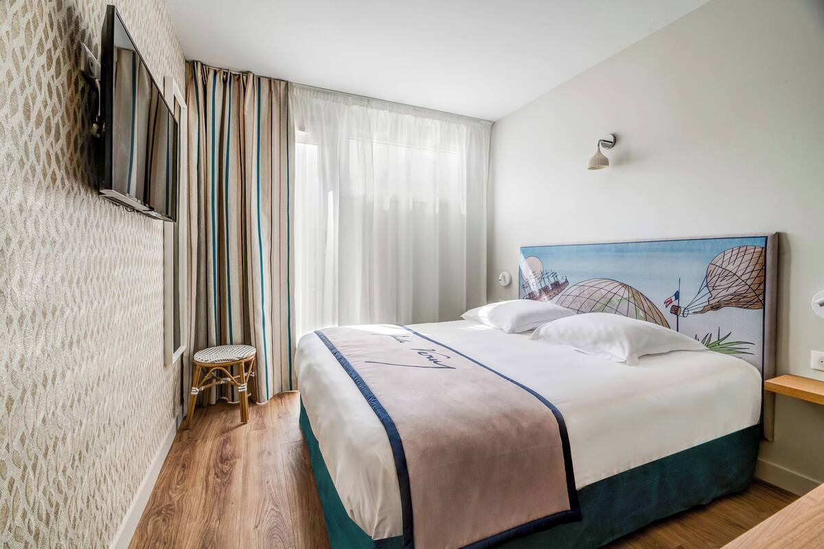 A hotel room with a bed, a small stool, a painting on the wall, and a TV mounted on the wall.