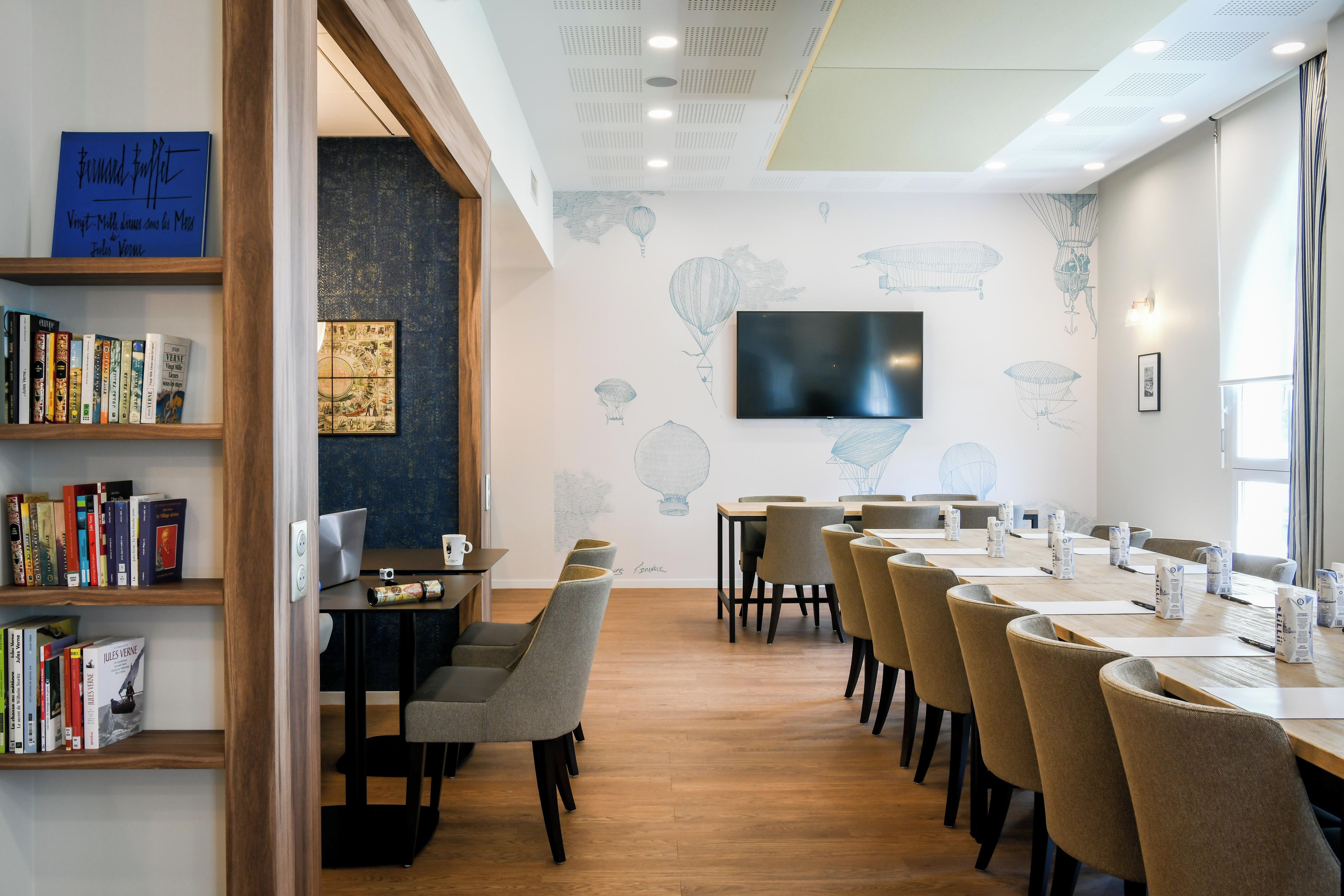 A modern conference room with a long table, chairs, a flat-screen TV, and bookshelves.