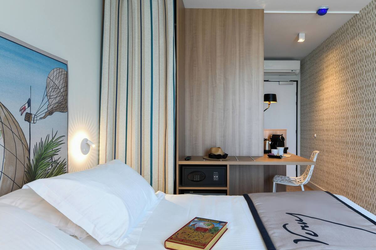 A hotel room with a bed, a desk, a chair, a lamp, and a wall-mounted air conditioner.