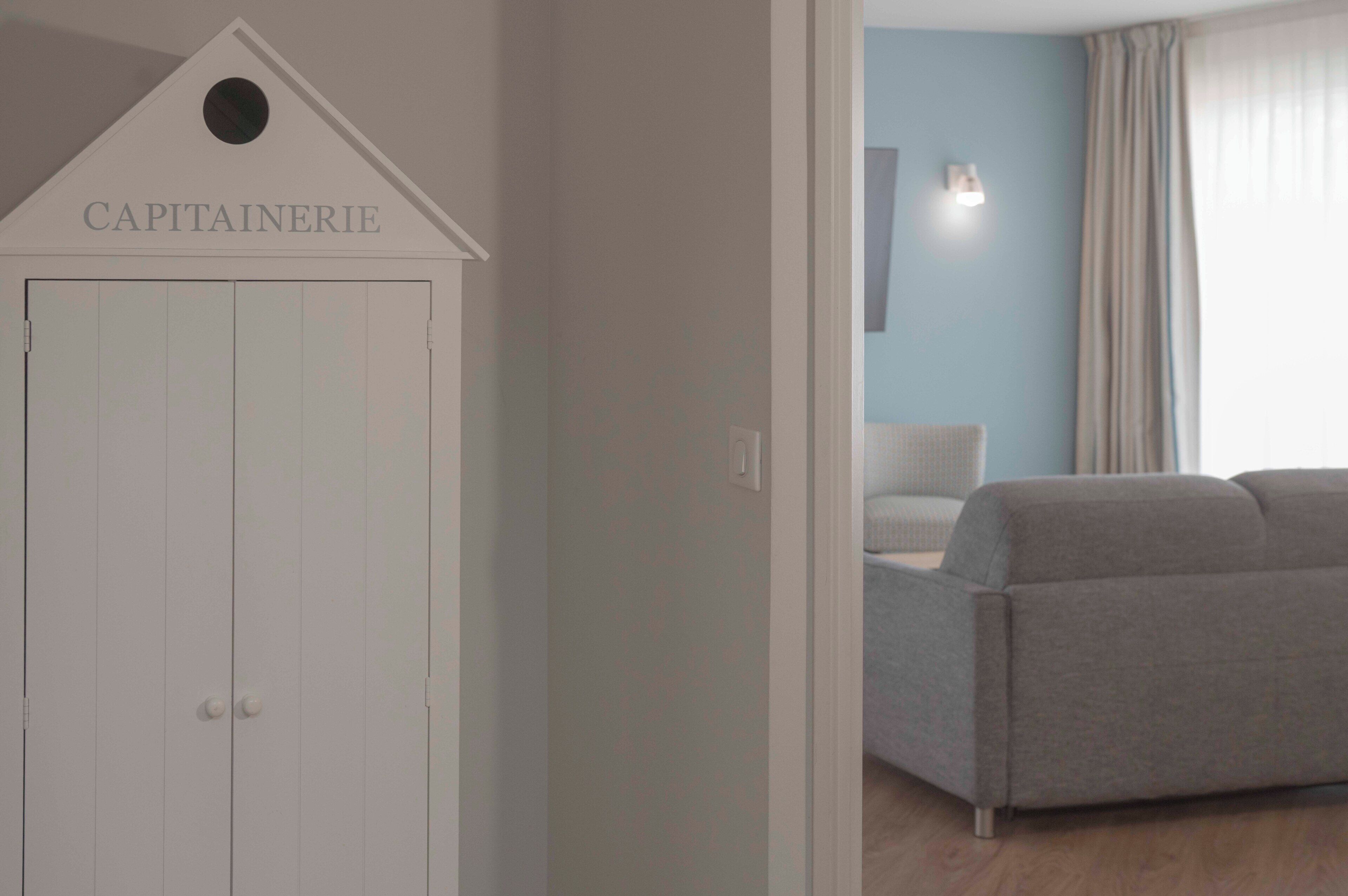 Ein Zimmer mit einem weißen Kleiderschrank mit der Aufschrift 'CAPITAINEERIE', einem grauen Sofa, einem Lichtschalter und einem mit Vorhängen versehenen Fenster.