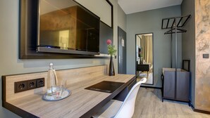 Una habitación de hotel moderna con un televisor de pantalla plana grande montado en la pared, un escritorio de madera con un vaso de agua y una botella, una silla blanca y un perchero.