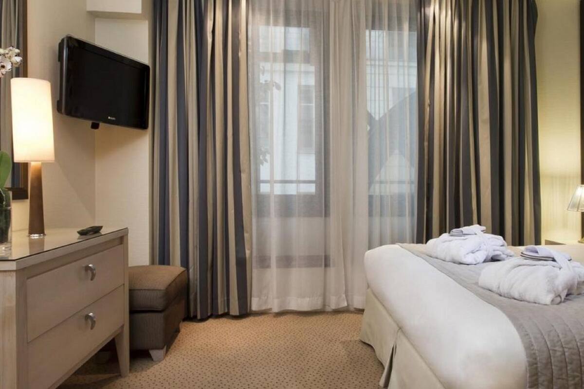 Une chambre d’hôtel avec un lit, un téléviseur fixé au mur, une table de chevet avec une lampe et une fenêtre avec des rideaux.
