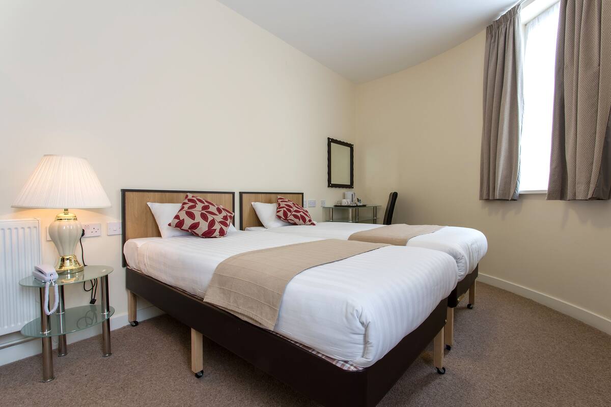 Une chambre d’hôtel avec deux lits, une table de chevet avec une lampe, une petite table avec un téléphone et une fenêtre avec des rideaux.