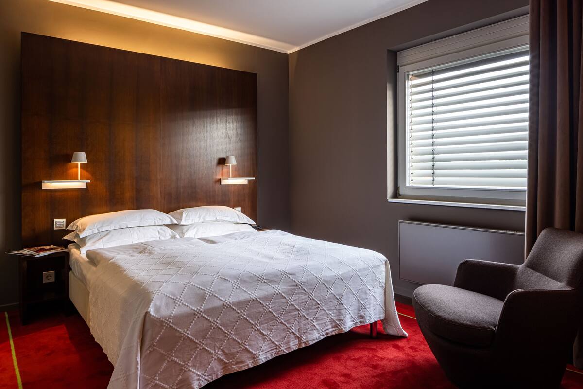 A hotel room with a large bed, two bedside lamps, a window with blinds, and a red carpet.