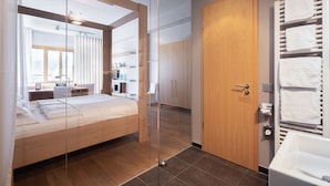 Una habitación de hotel moderna con un separador de vidrio, suelo de madera, una cama, un escritorio, un armario y un baño con bañera y tendedero para toallas.