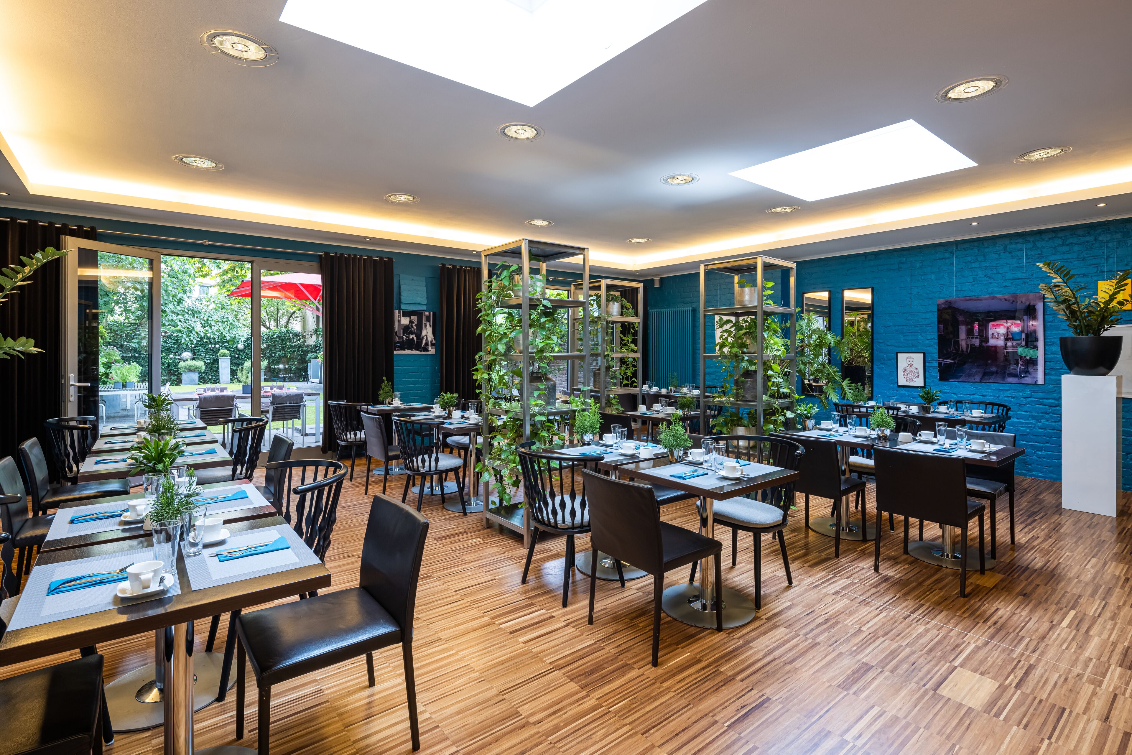 Ein modernes Restaurant mit Holzboden, mehreren Essplätzen, schwarzen Stühlen und Topfpflanzen.