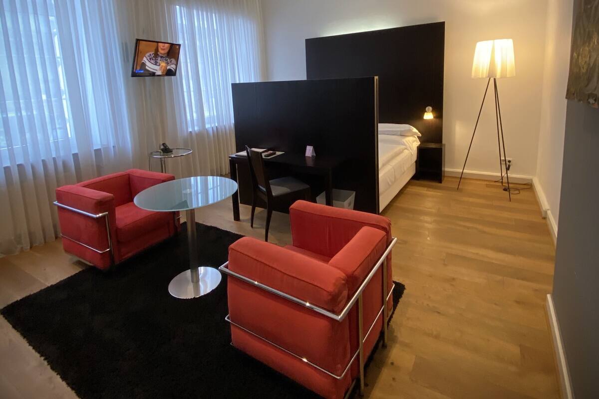 A modern hotel room with a bed, a desk, two red armchairs, a small round table, and a TV mounted on the wall.