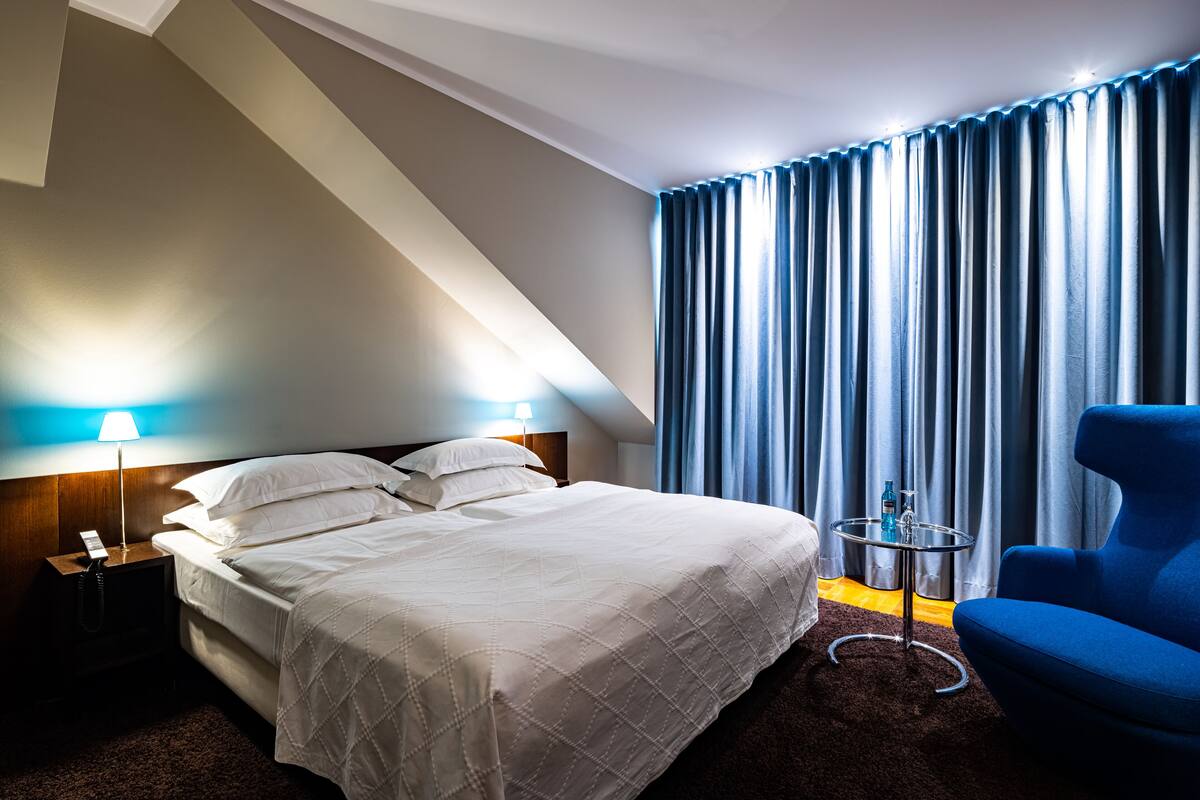 A hotel room with a bed, a nightstand with a lamp, a blue chair, and a small table with a bottle.