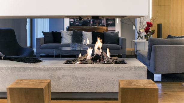 Un salón moderno con una chimenea central, sofás grises y suelo de madera.