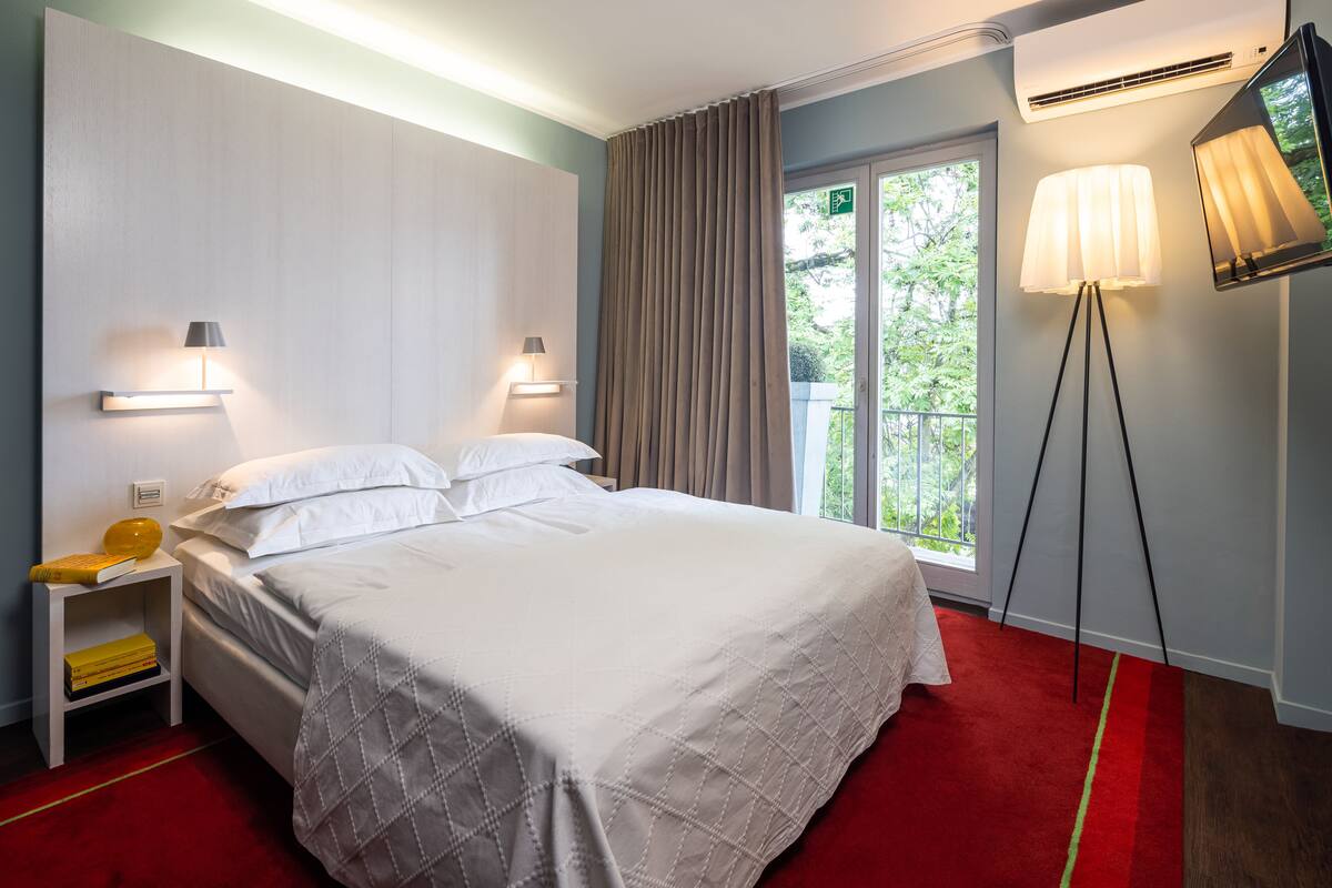 A hotel room with a large bed, bedside lamps, a nightstand with a book, a red carpet, and a window with curtains.