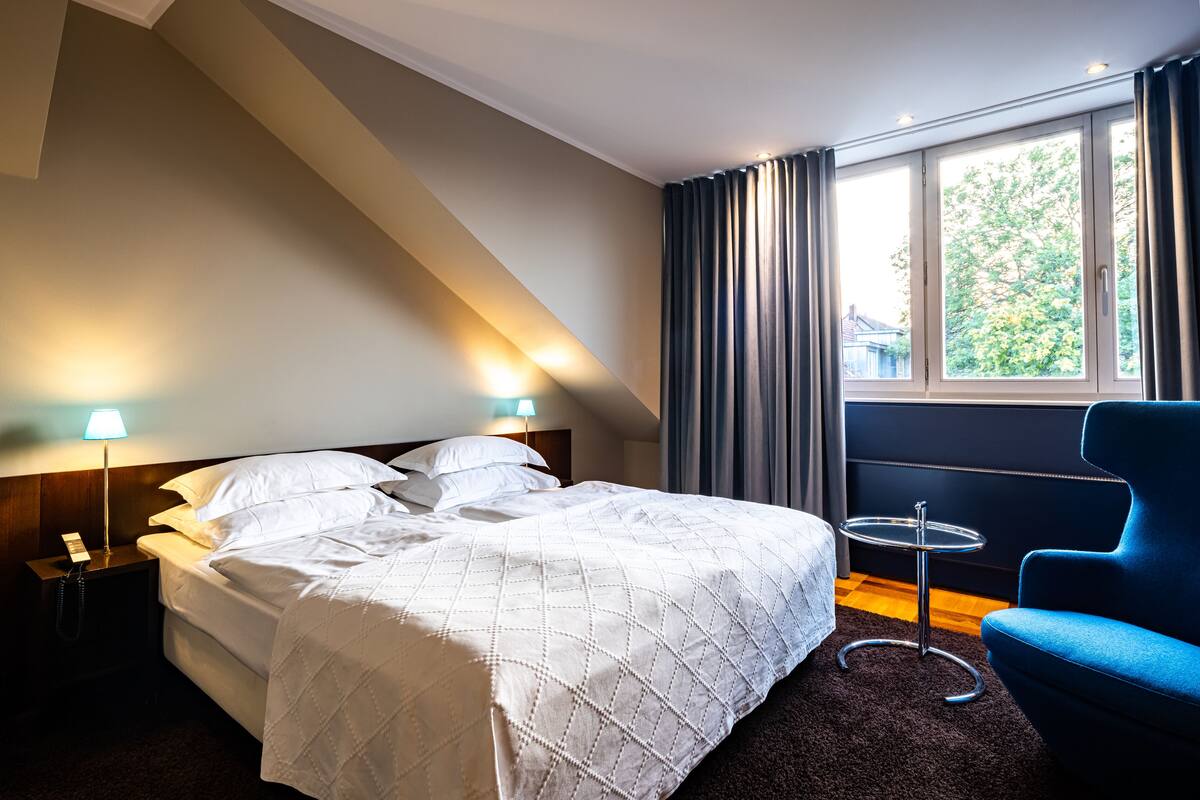 A hotel room with a bed, a nightstand, a blue armchair, a small round table, and a window with curtains.