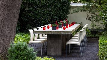 Una larga mesa de comedor con sillas, rodeada de follaje y un árbol.