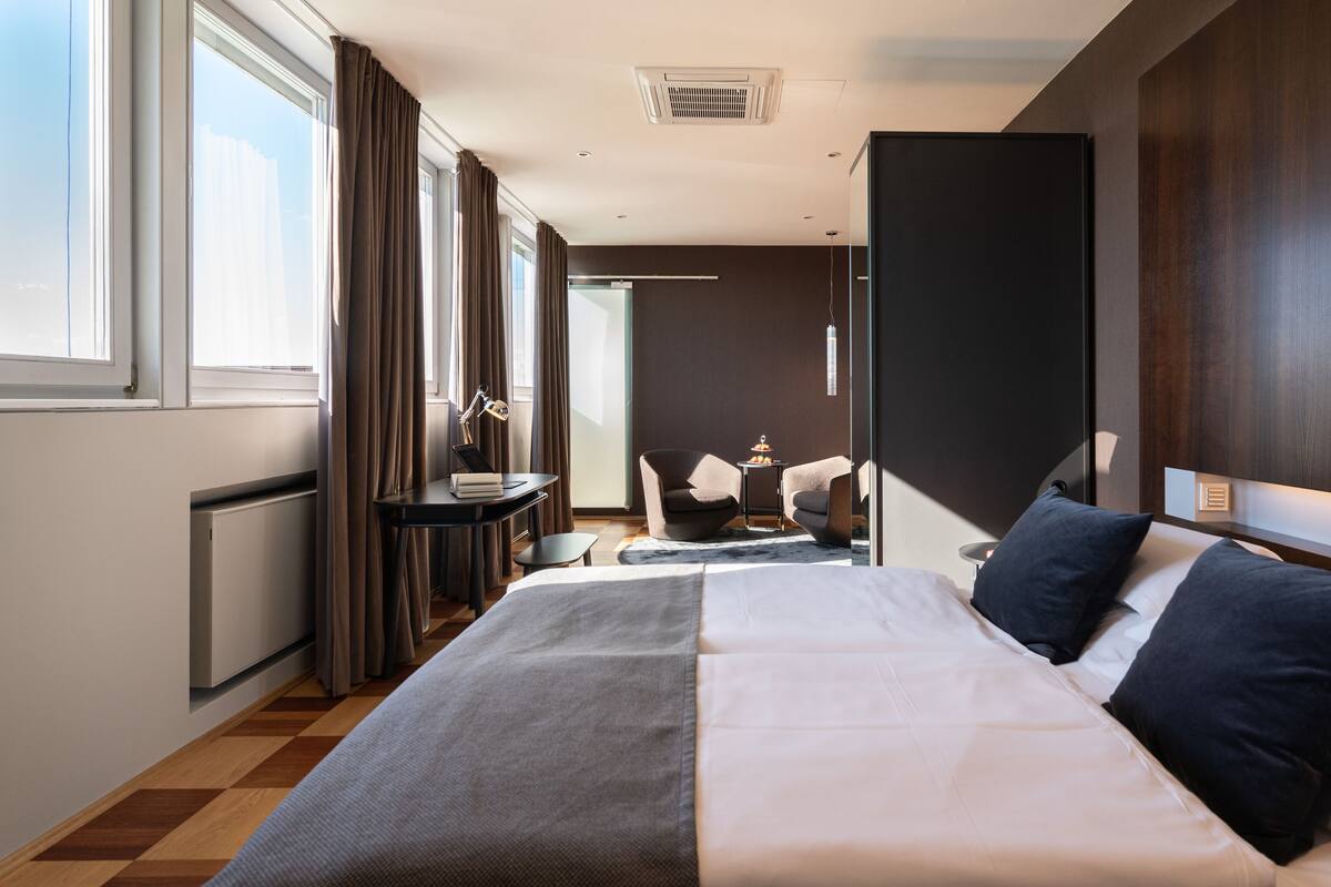 A modern hotel room with a large bed, a desk with a chair, a small table with a lamp, and a view of the city.