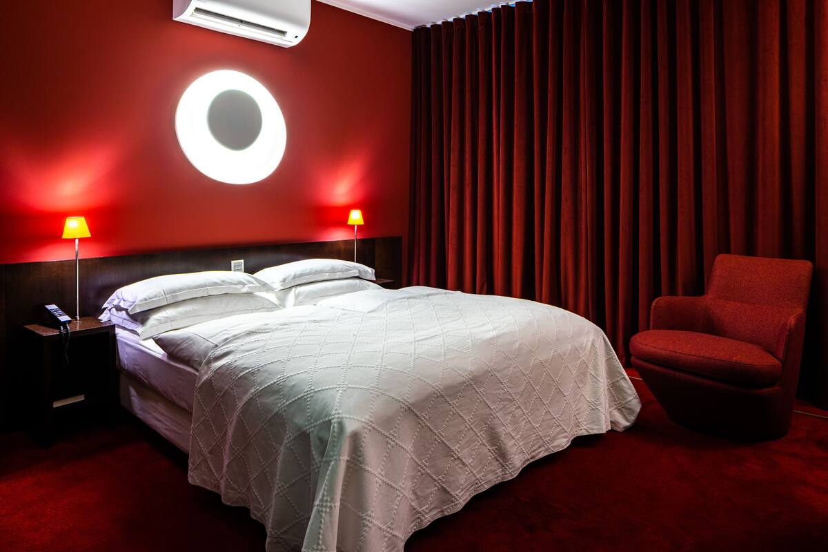 A hotel room with a large bed, two bedside lamps, a red chair, and a circular light fixture on the wall.