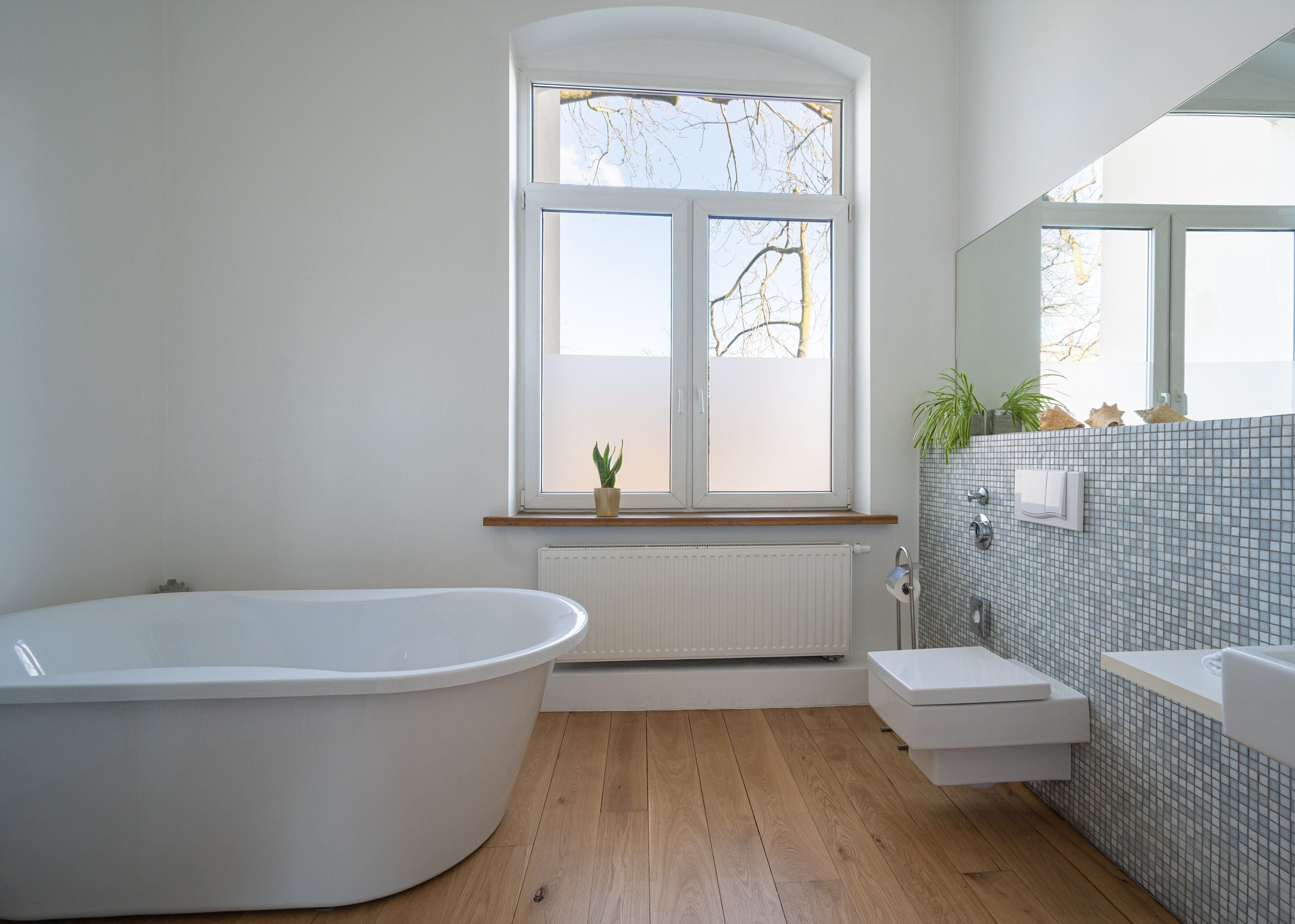 Un baño moderno con una bañera independiente, un lavamanos pequeño y una ventana con vistas a árboles desprovistos de hojas.