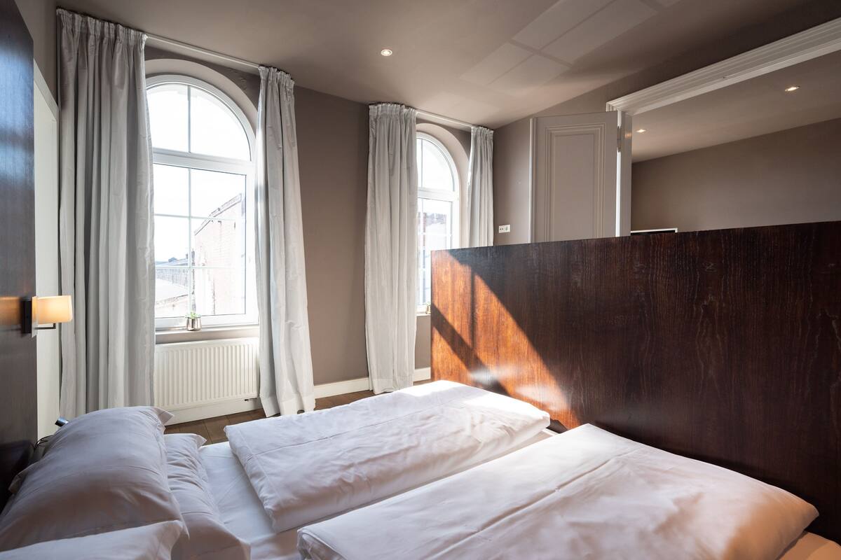 A hotel room with two beds, large windows with curtains, a wooden headboard, and a wall-mounted light.
