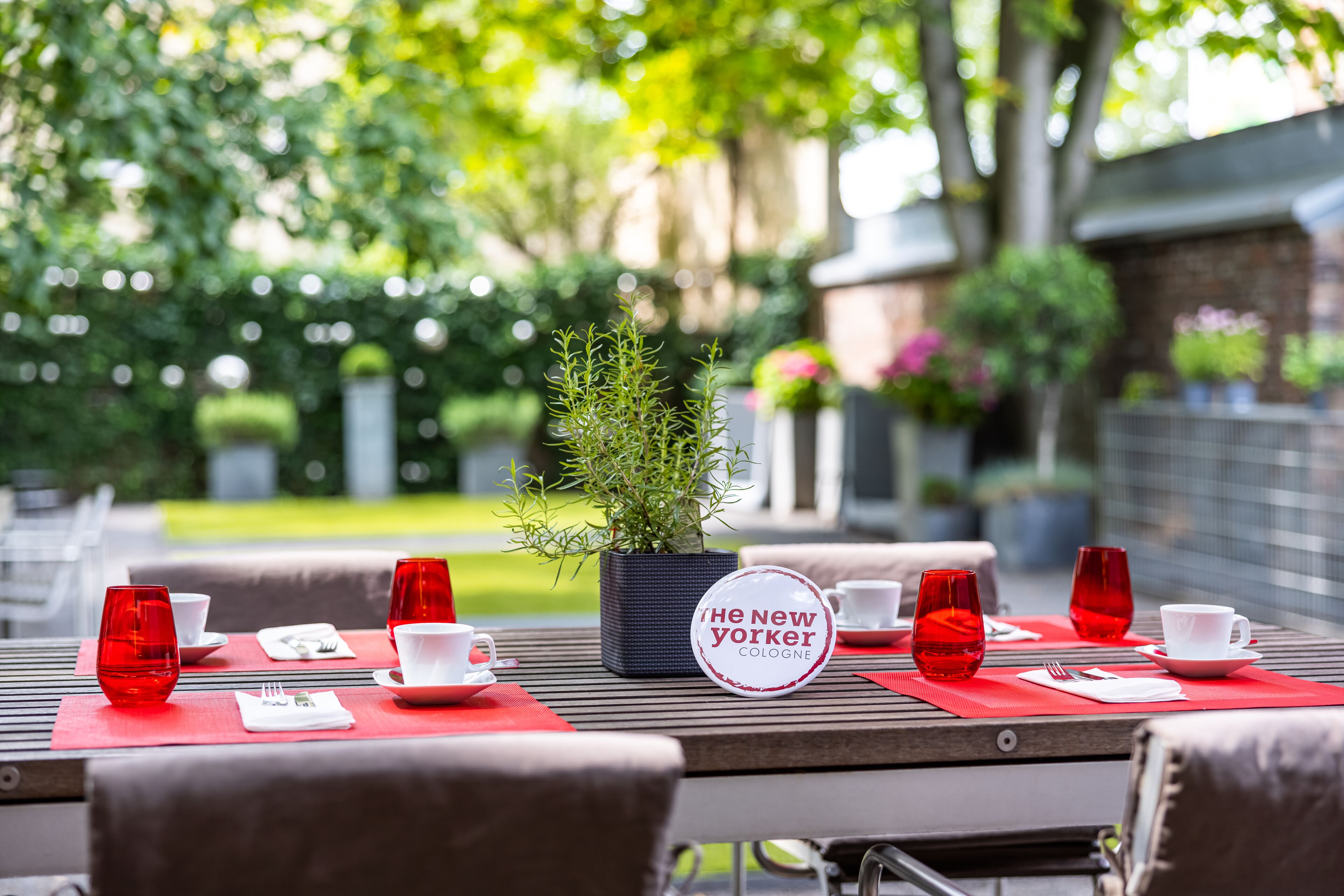 Un espacio de comedor al aire libre con una mesa preparada para dos personas, con individuales rojos, tazas blancas y un letrero que dice "The New Yorker Cologne".