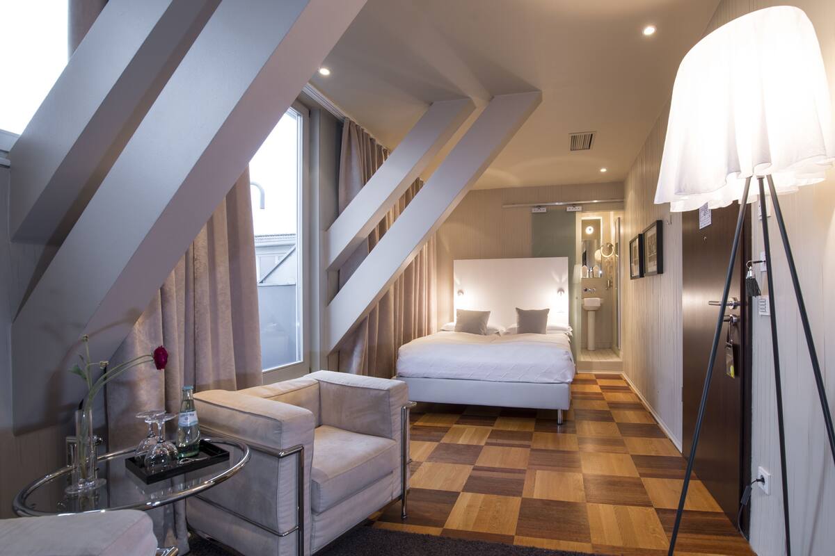 A modern hotel room with a checkered floor, a bed, a sitting area with a table and chair, and a large window.