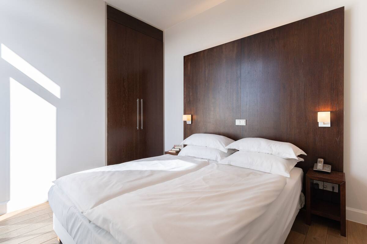 A modern bedroom with a large bed, wooden headboard, bedside lamps, and a nightstand.