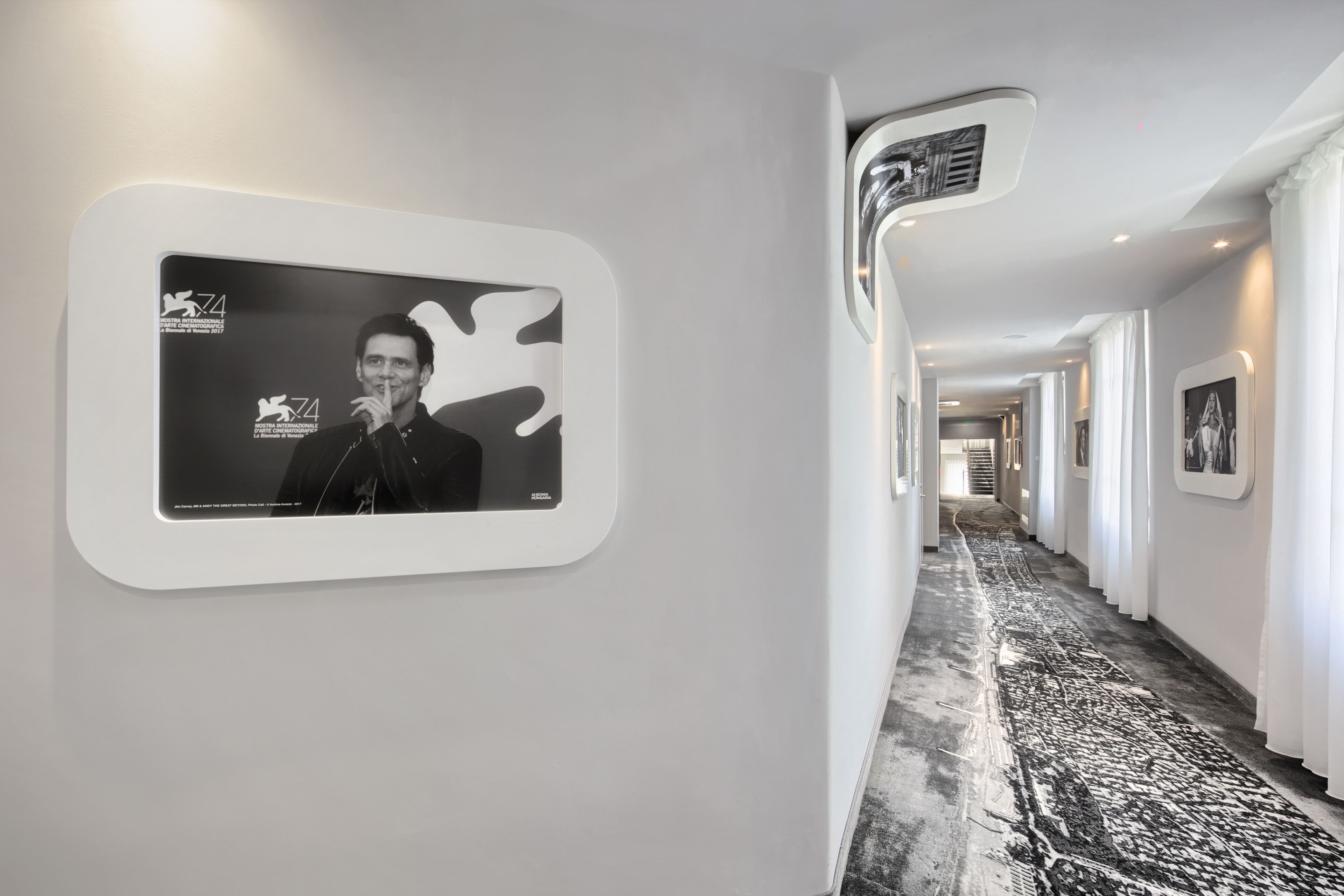 A hallway with a patterned carpet, framed artwork on the walls, and a mounted screen displaying a person.