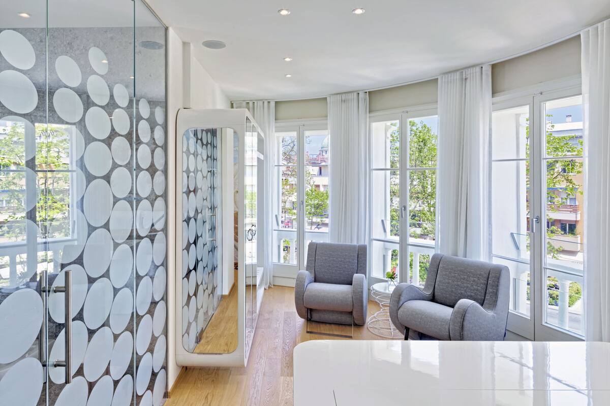A modern hotel room with large windows, a glass partition with a circular pattern, and a seating area with two armchairs.