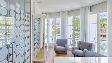 A modern hotel room with large windows, a glass partition with a circular pattern, and a seating area with two armchairs.