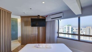 Une chambre d’hôtel moderne dotée d’un grand lit, d’un téléviseur à écran plat fixé au mur et offrant une vue sur la ville grâce à de grandes fenêtres.