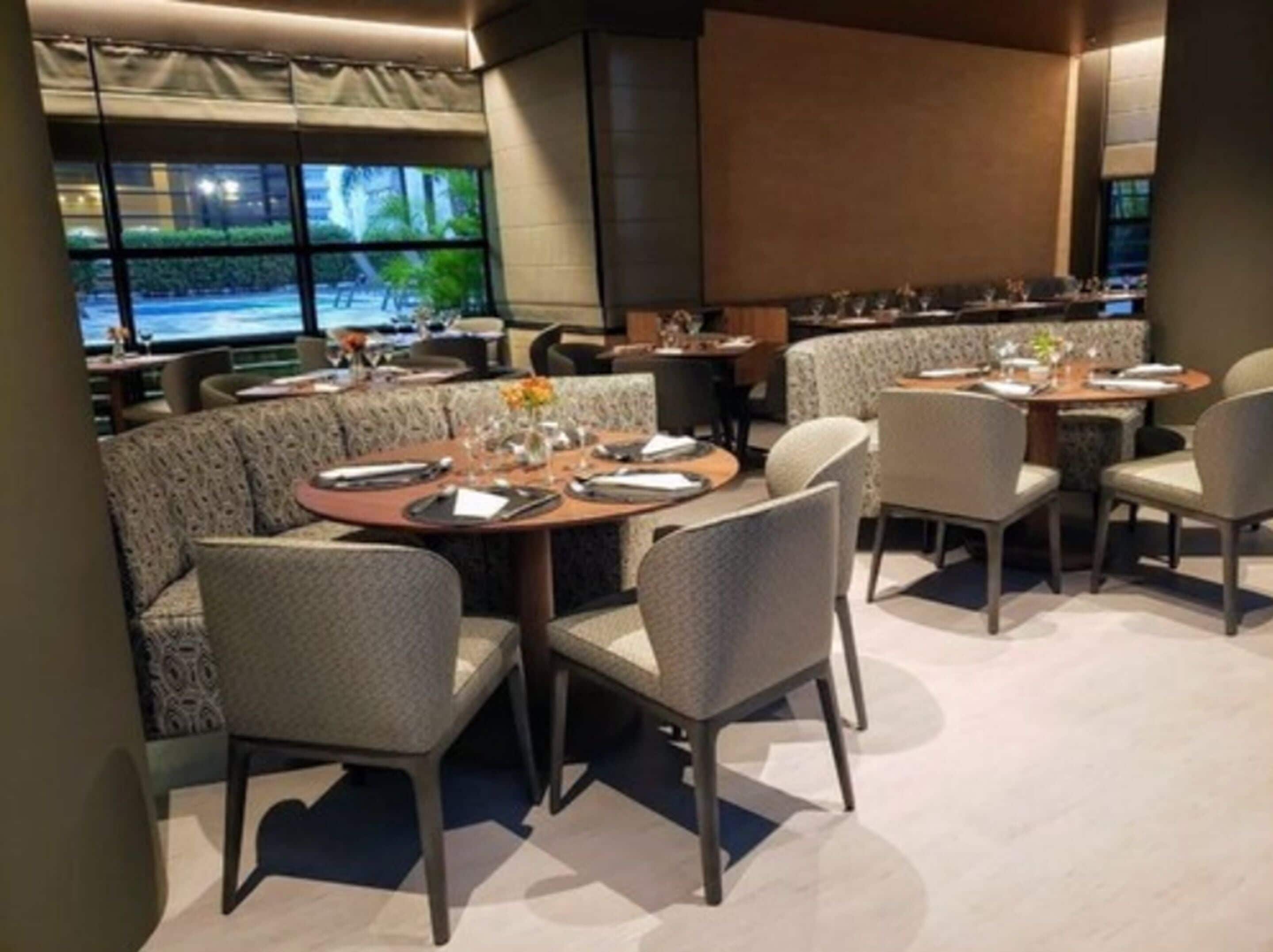 A restaurant with round tables and cushioned booths, large windows with a view of palm trees, and a modern interior design.