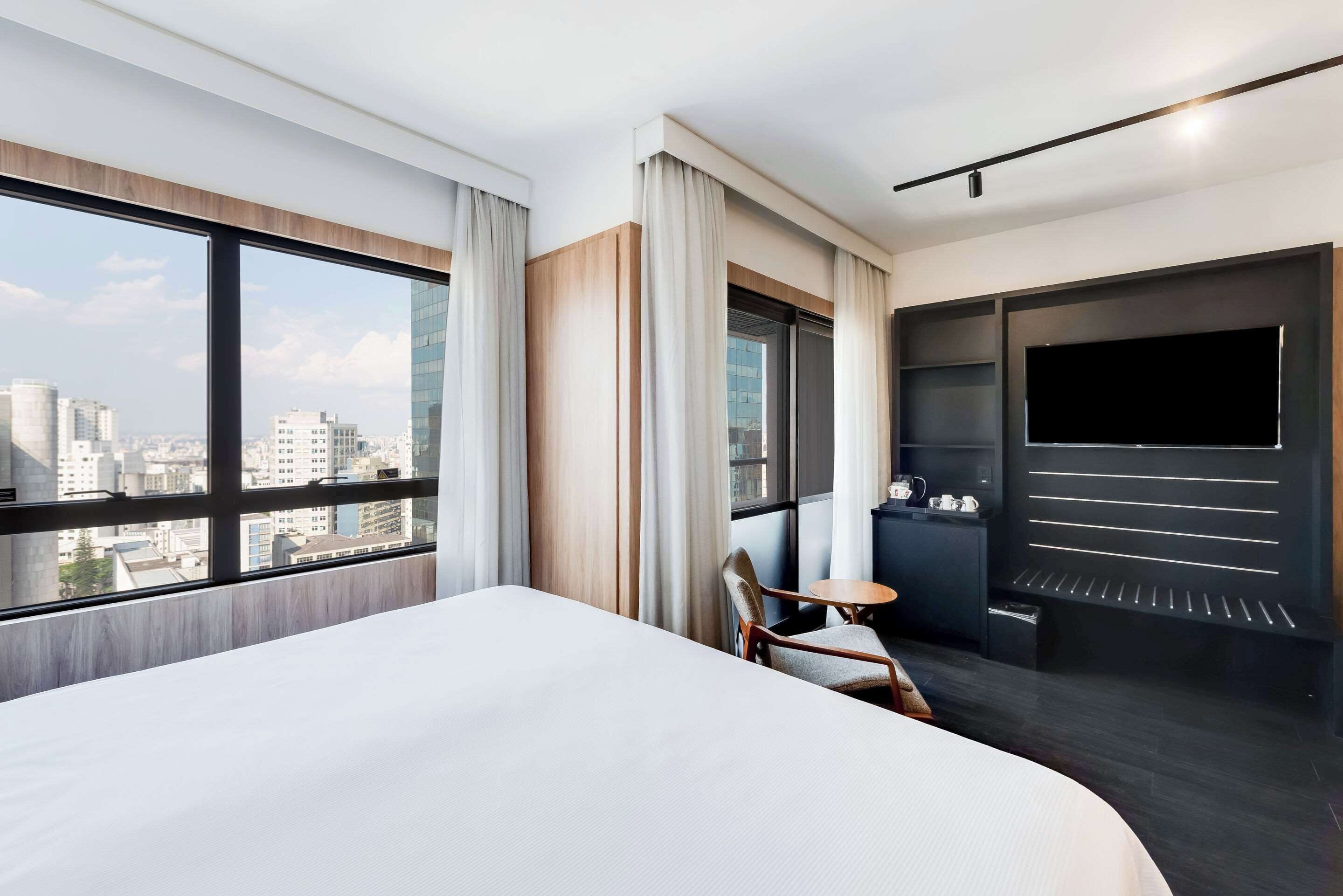 Quarto de hotel com uma cama grande, televisão, uma mesinha e uma cadeira.