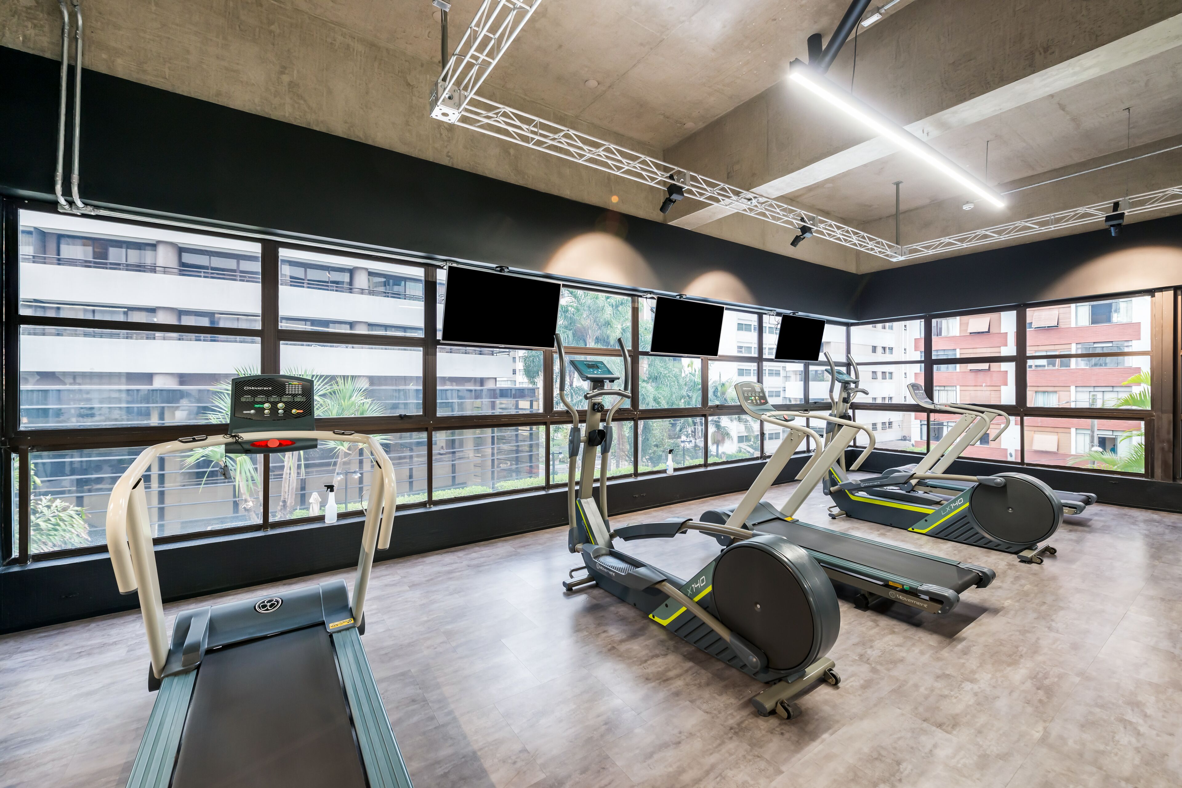 A gym with treadmills, elliptical machines, and stationary bikes. Large windows provide views of buildings outside.