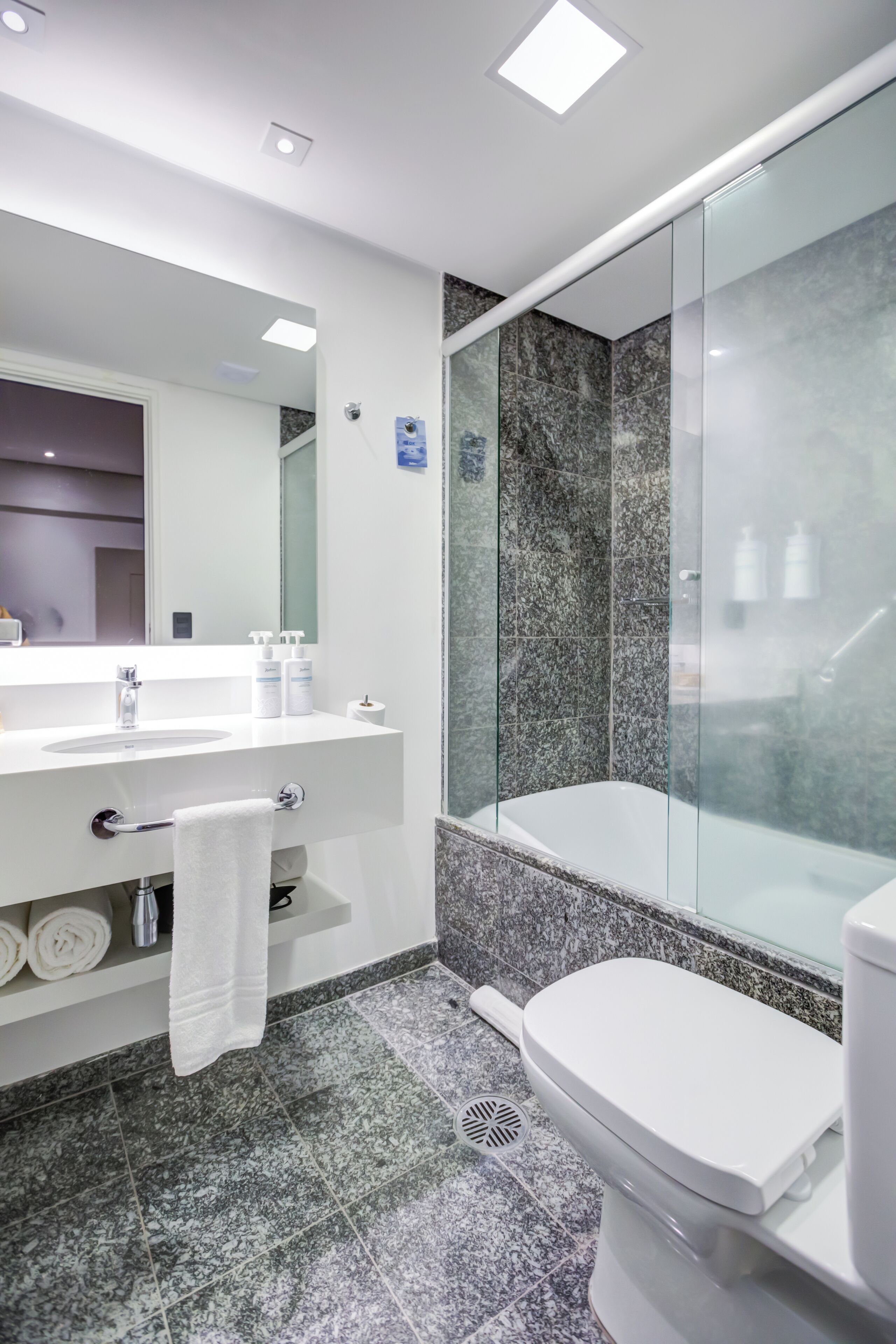 Un baño moderno con un lavamanos blanco, espejo y inodoro, además de una zona de ducha con mampara de vidrio.