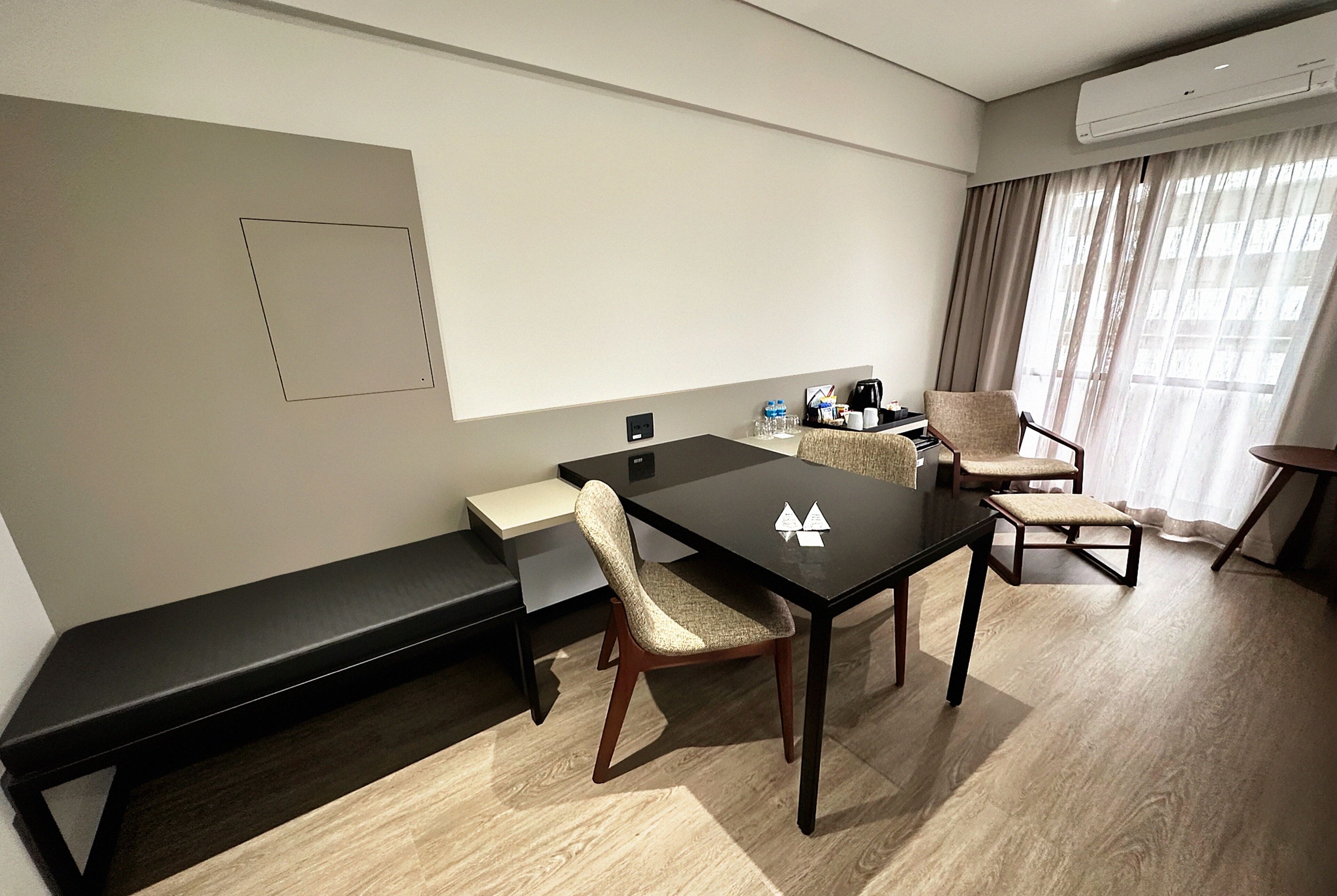 Ett modernt hotellrum med trägolv, ett svart matbord, två stolar, en bänk, ett litet bord med en kaffebryggare och ett fönster med gardiner.
