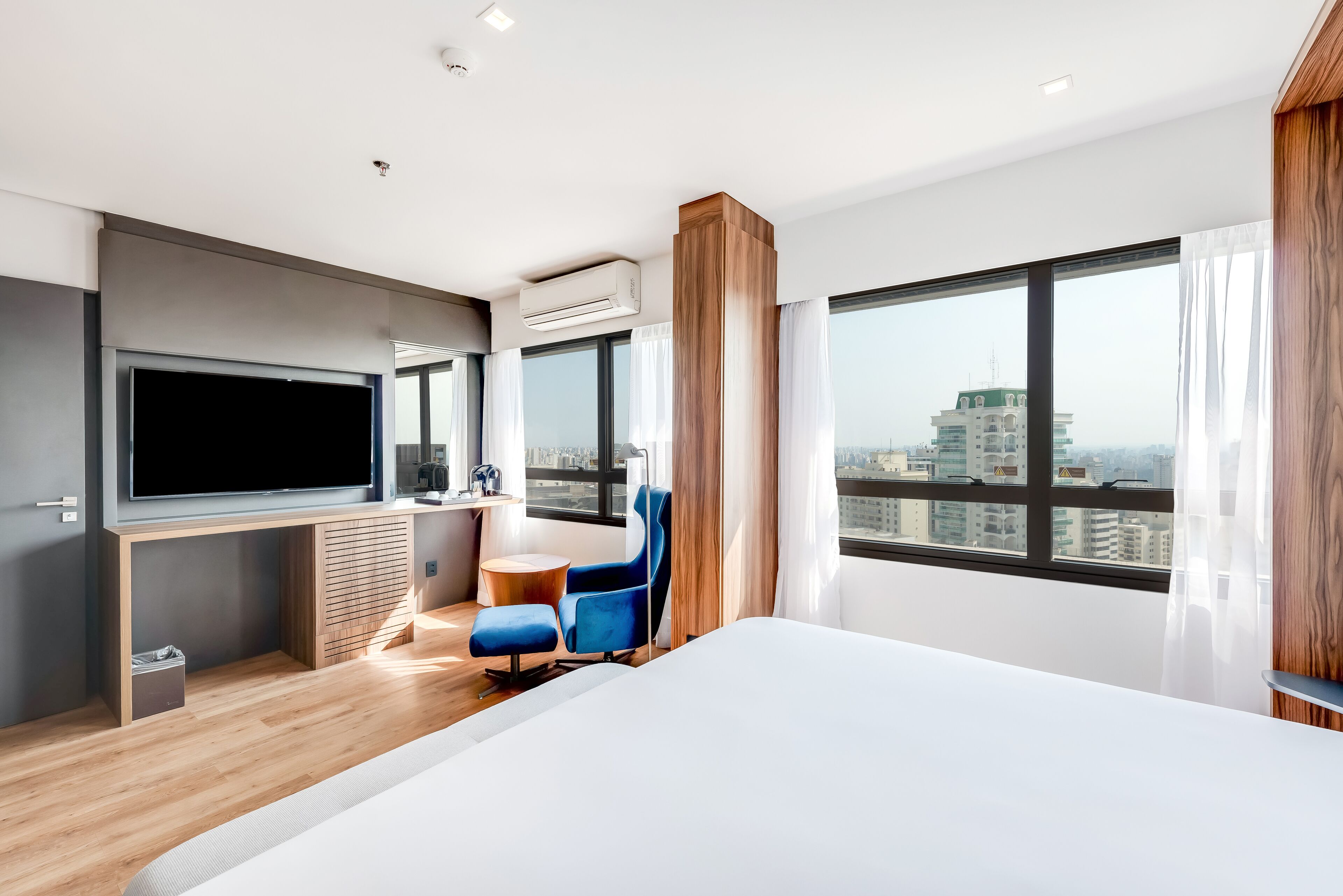 A modern hotel room with a large bed, a flat-screen TV, a desk, a blue chair, and a city view through large windows.