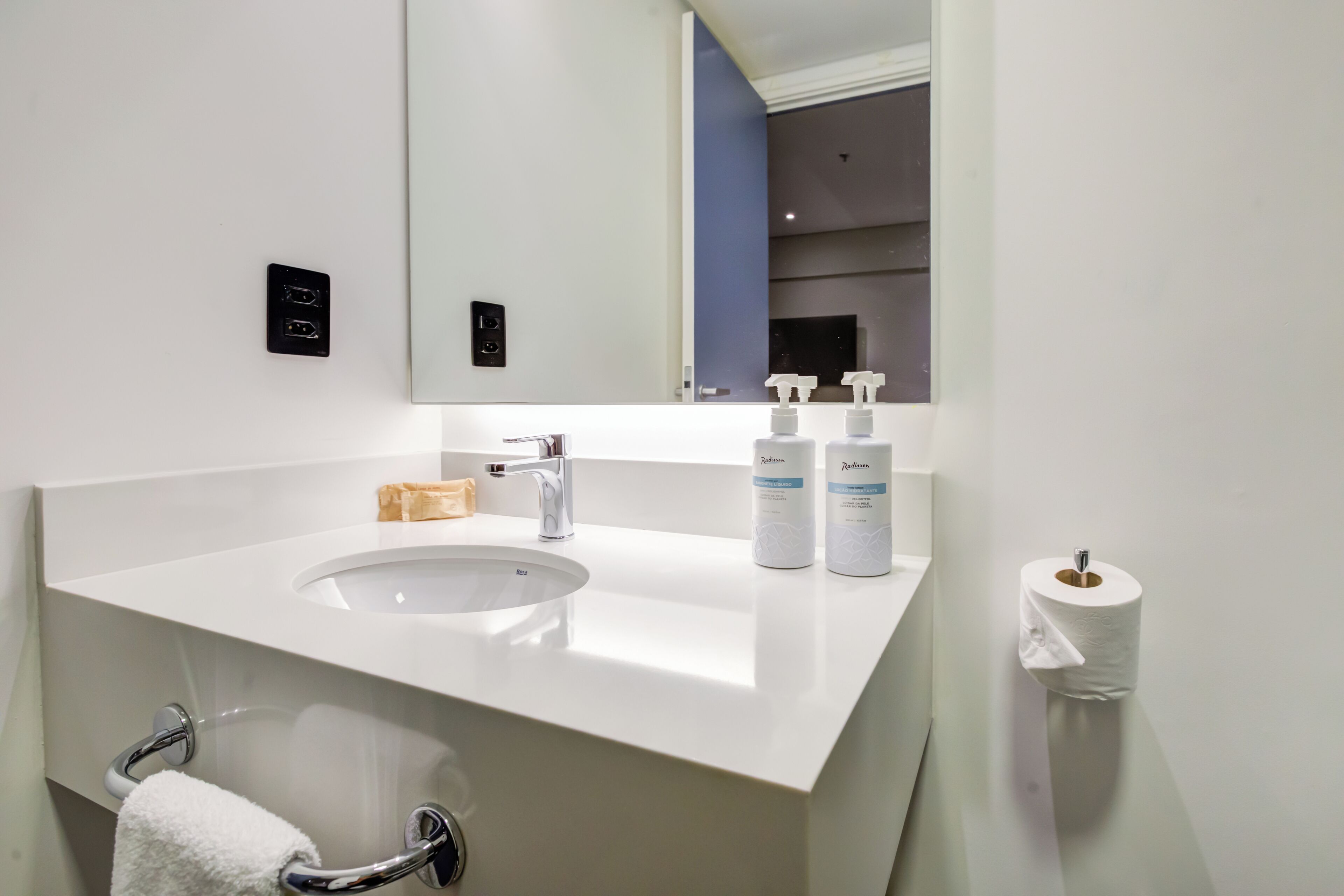 A modern bathroom with a white sink, a mirror, a towel rack, and two bottles of Radisson hand soap.