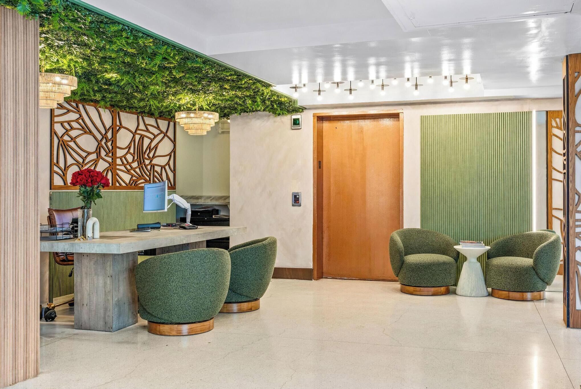 A modern reception area with a reception desk, green upholstered chairs, a small side table, and a wooden door.