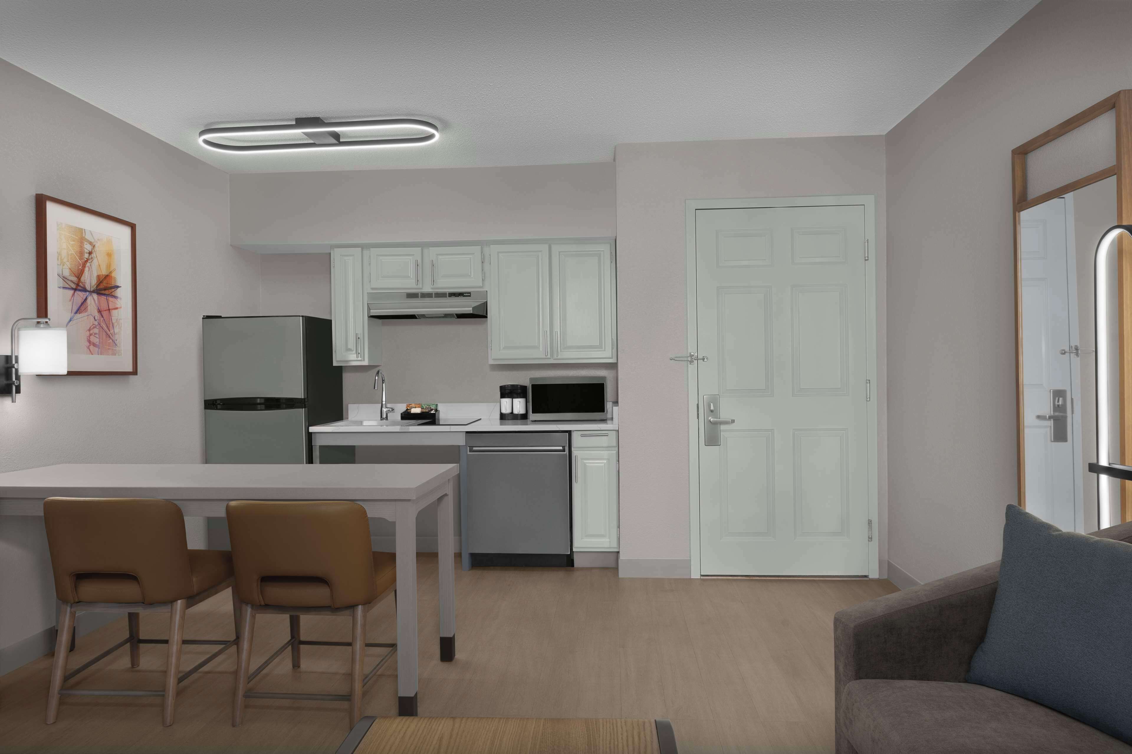 A modern kitchen with white cabinets, a refrigerator, microwave, and a coffee maker. There is a dining table with two chairs and a sofa in the living area.