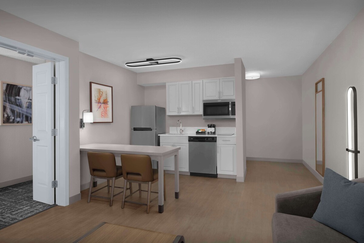 A modern kitchen with white cabinets, a stainless steel refrigerator, and a microwave. There is a dining area with a white table and two chairs.