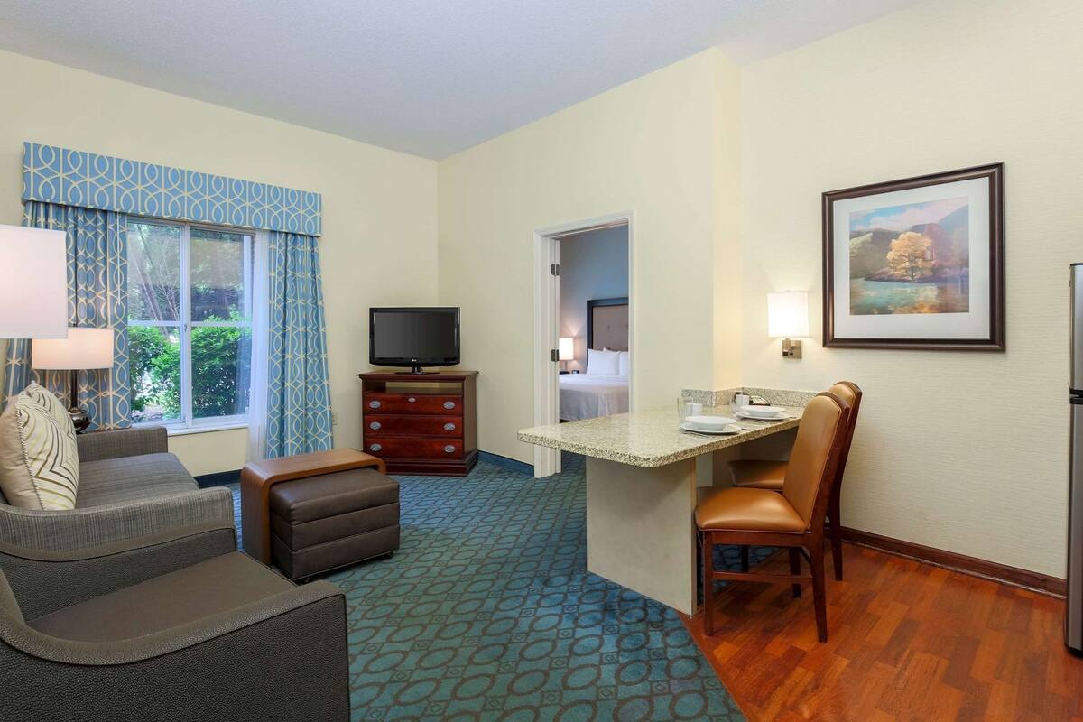 A hotel room with a sofa, ottoman, TV, dresser, dining table, and chairs.
