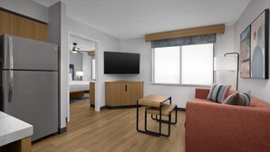 A modern, well-lit room with a kitchen area, a sofa, a TV, and a window with blinds. - Homewood Suites by Hilton Ft. Worth-North at Fossil Creek (Fort Worth)
