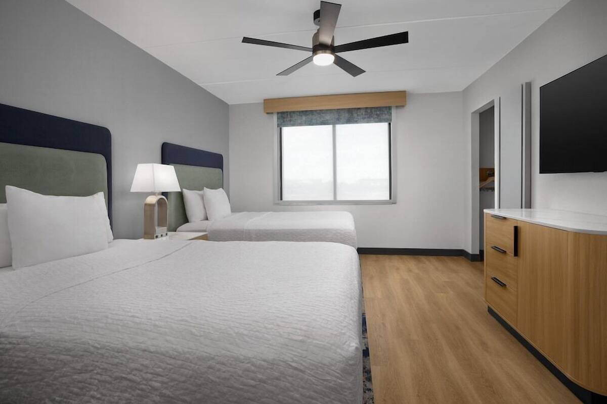 A hotel room with two beds, a ceiling fan, a large window, a TV, and a wooden cabinet.