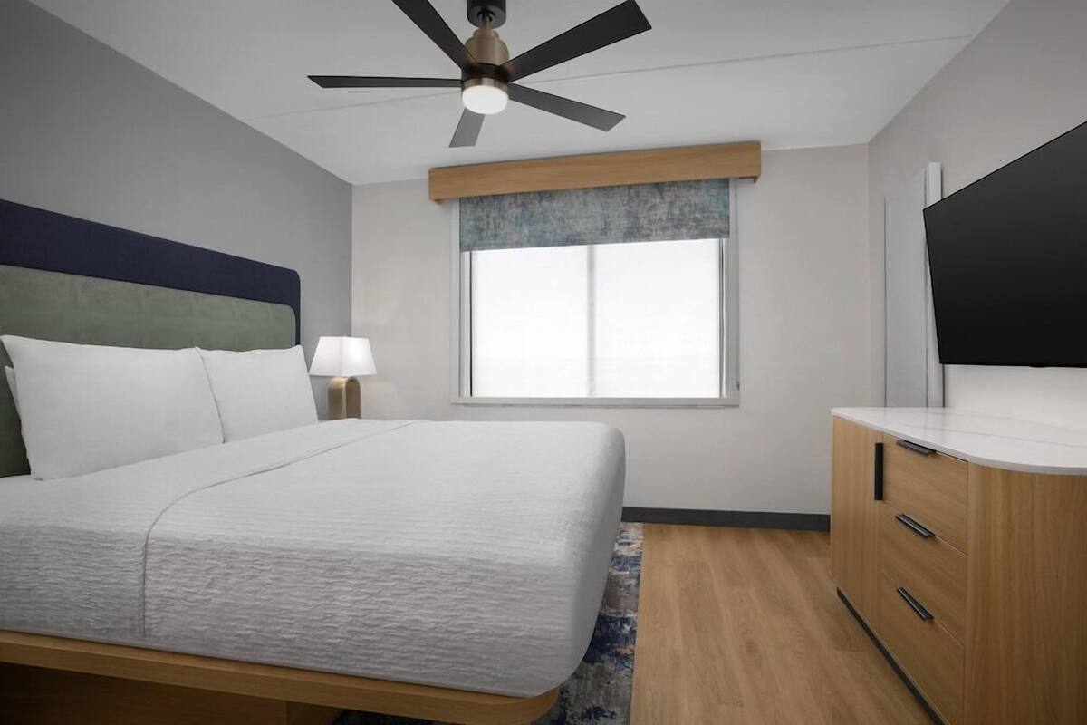 A hotel room with a bed, a ceiling fan, a window with blinds, a TV, and a wooden dresser.