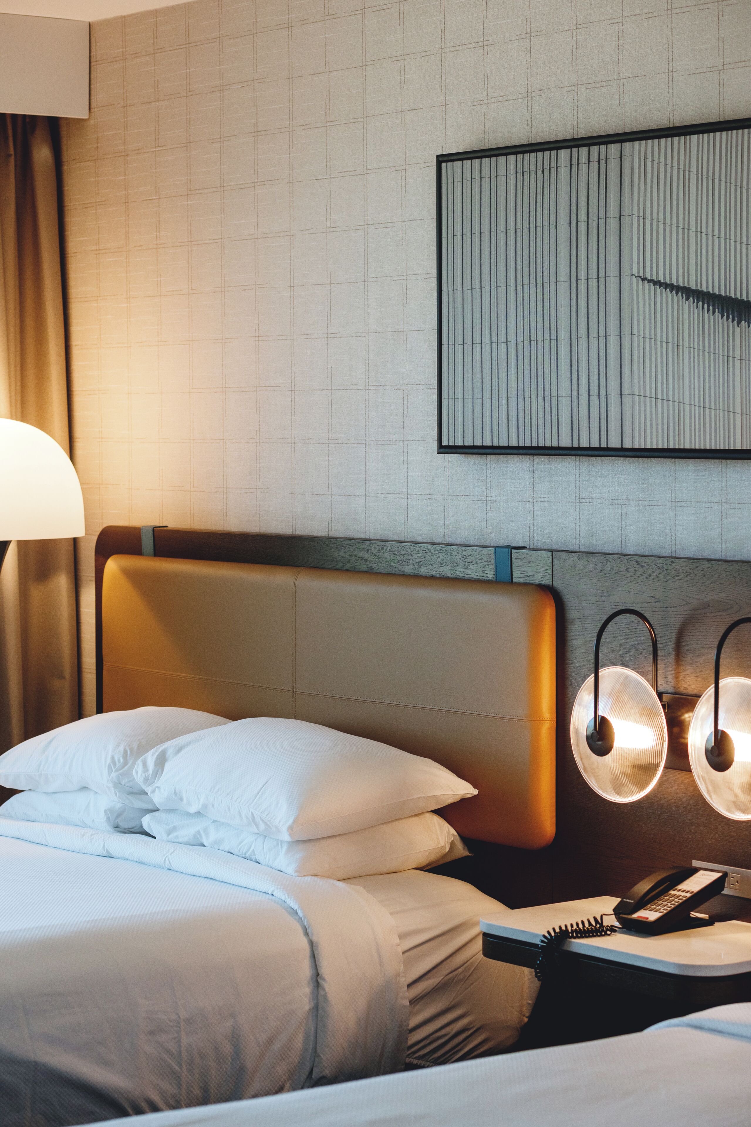A hotel room with a bed, a bedside table with a lamp, a phone, and a wall-mounted light fixture.